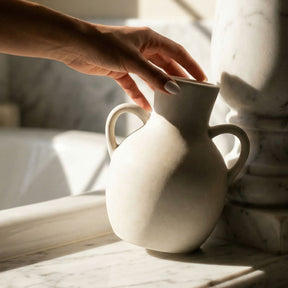 Person holding a white ceramic vase on a marble surface with soft lighting