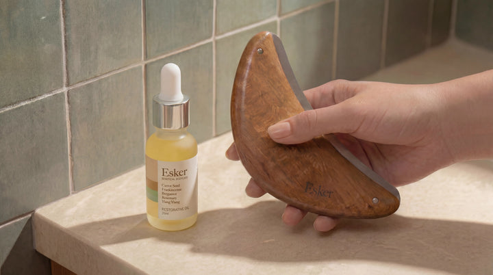 Esker Restorative Oil Deluxe Mini beside a hand holding a wooden gua sha tool