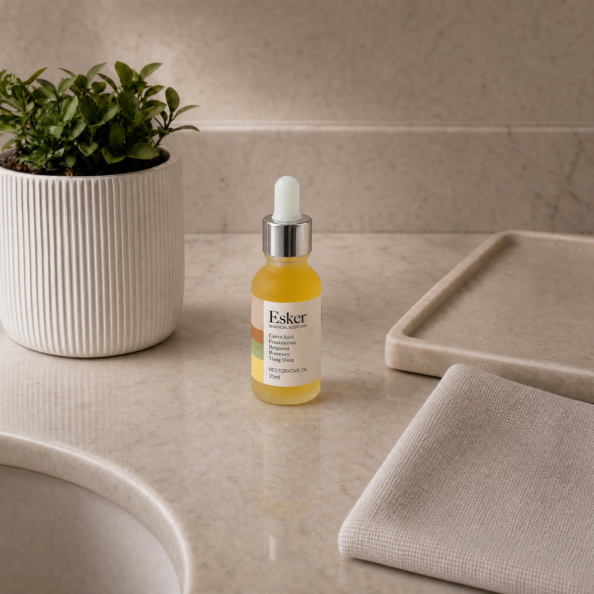 Esker Restorative Oil Deluxe Mini on a marble counter with folded towels and a plant