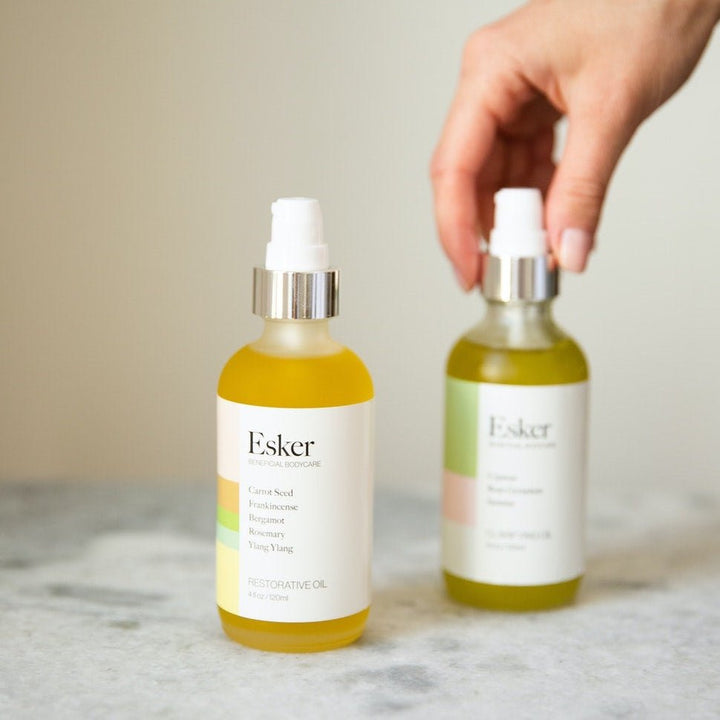 Hand reaching for one of two Esker Restorative Body Oil bottles on marble